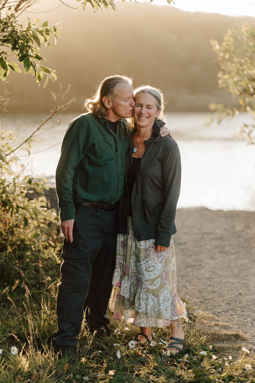 A couple shares a tender kiss by a sunlit river, the man in green and woman in a floral skirt smiling warmly.