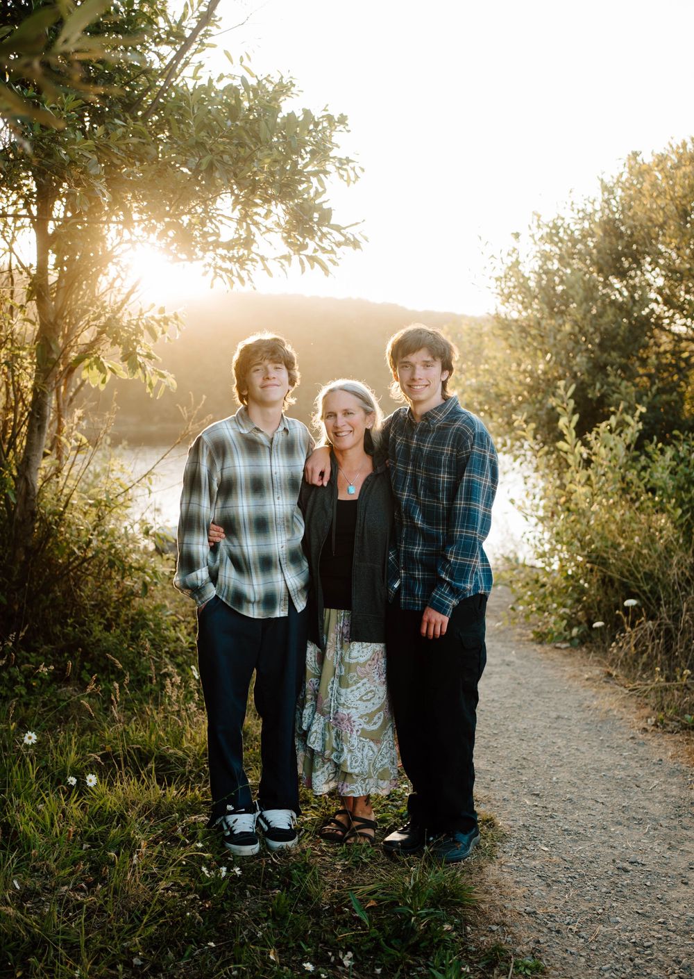A woman and two teenage boys pose together on a sunlit nature path beside a lake during golden hour.