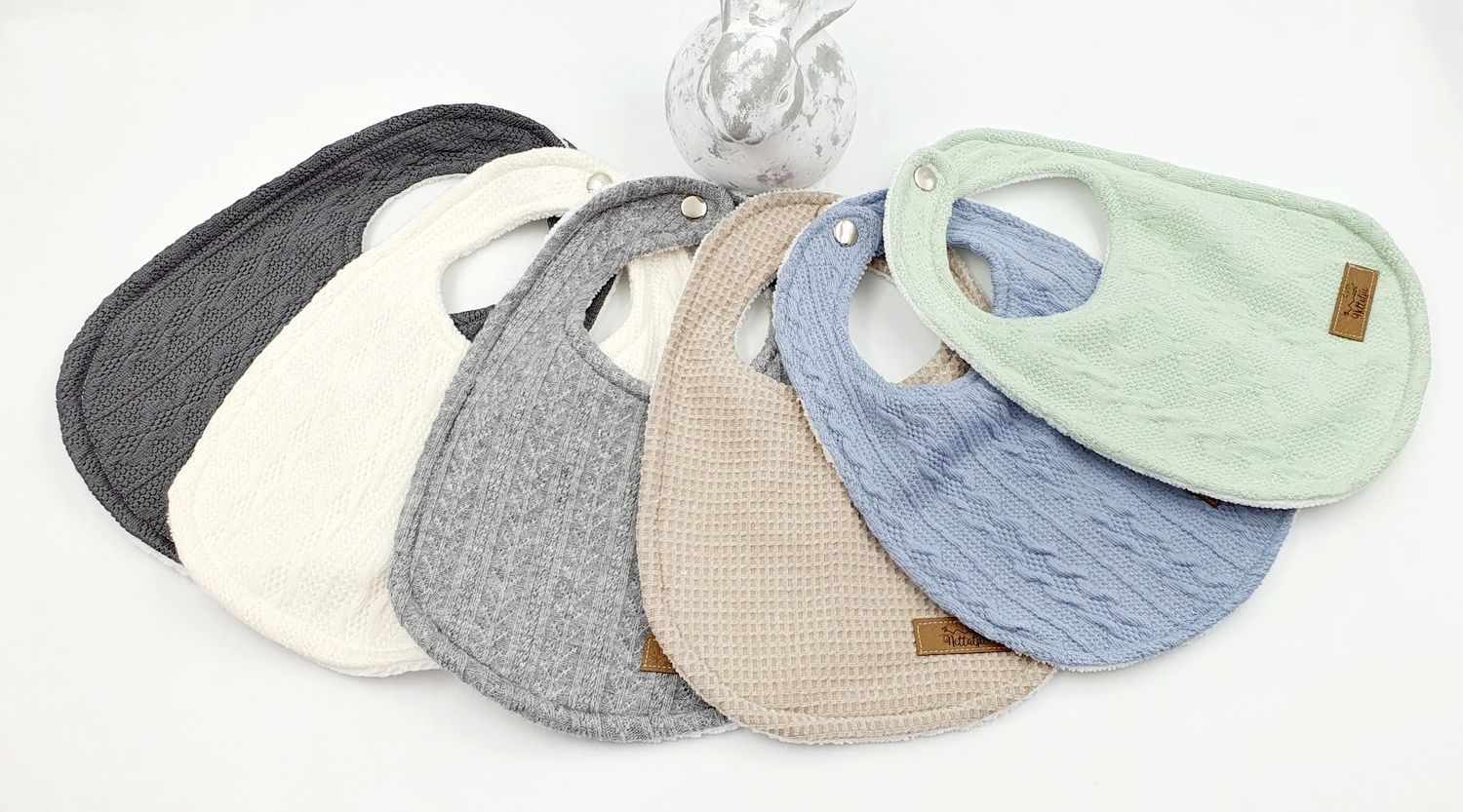 Set of seven colorful baby bibs in various textures including knit and waffle patterns in neutral and pastel tones.