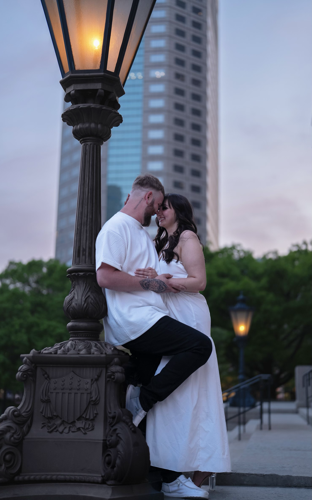 Romantic couple embracing near an ornate street lamp at dusk with city skyscraper backdrop.