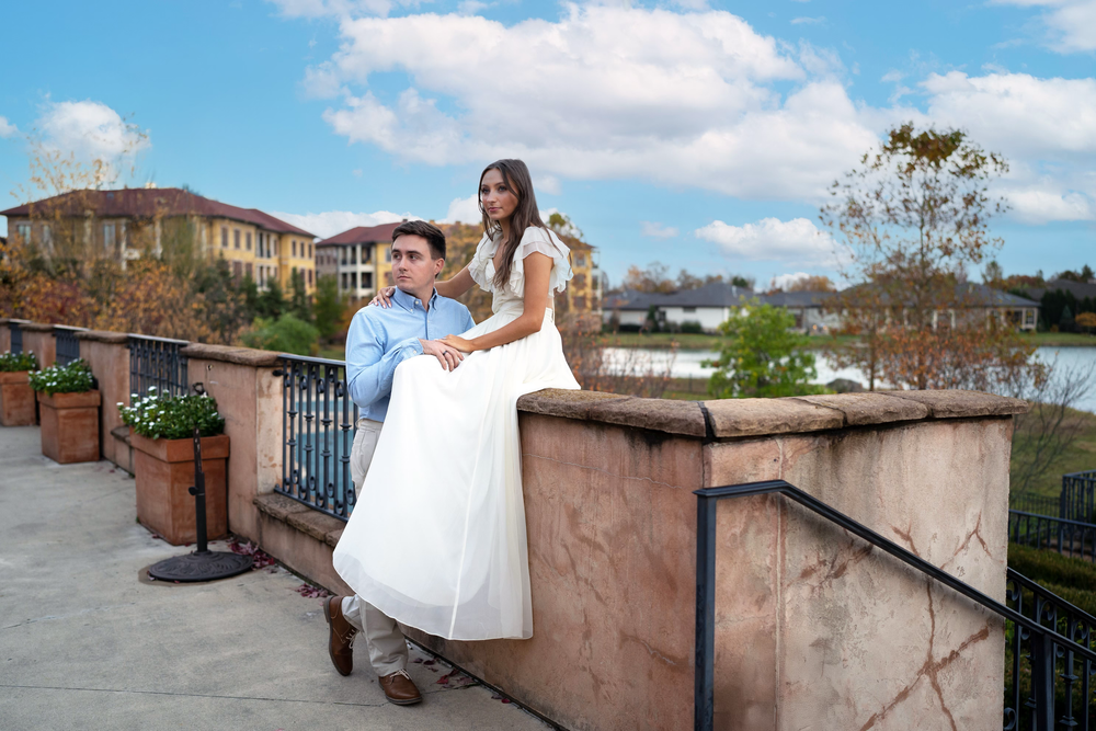 Couple posing on a terrace balcony near a lake, woman in white dress, man in blue shirt, on a sunny day.