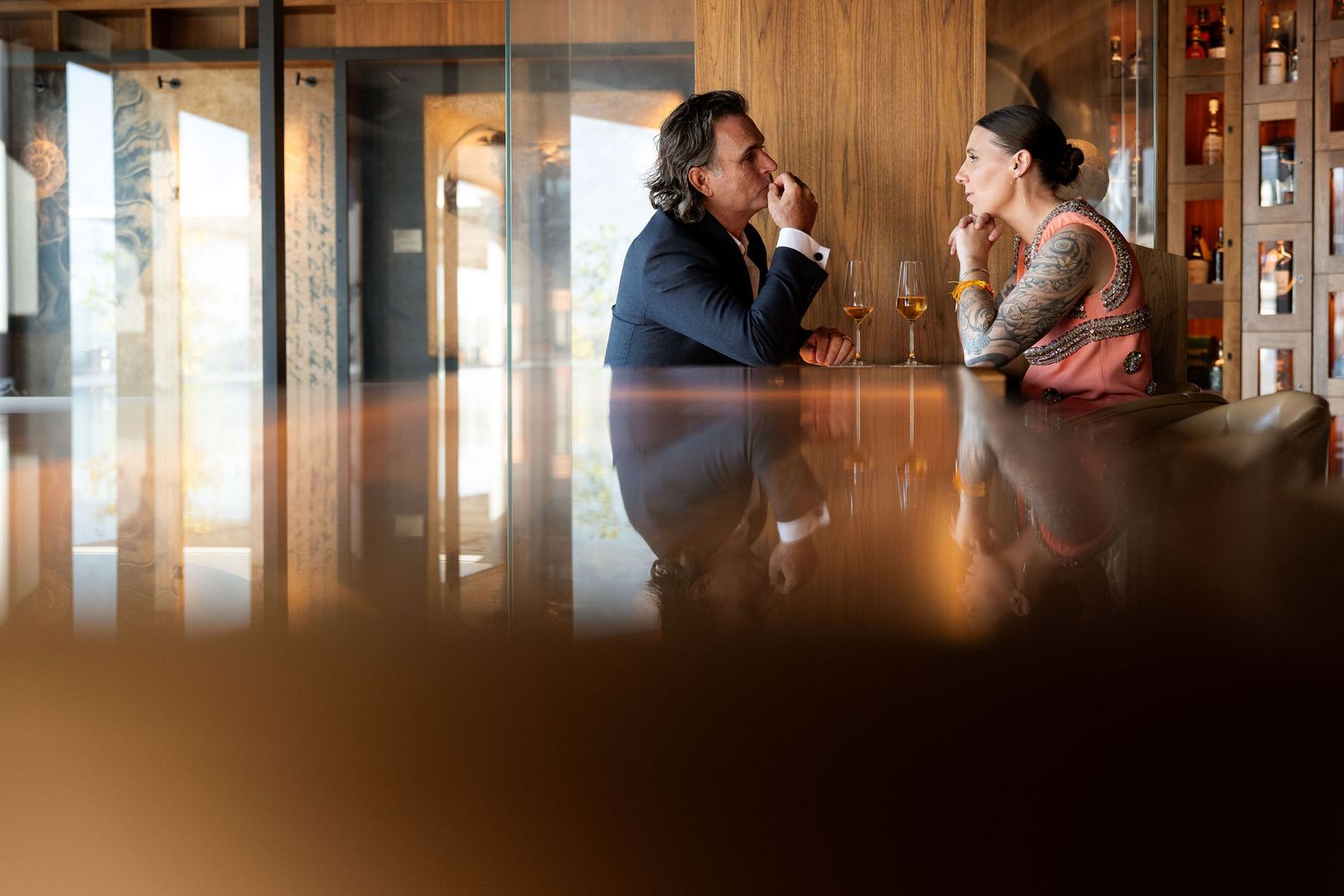 A man and woman engaged in conversation at a sophisticated bar, seated across a reflective wooden counter.