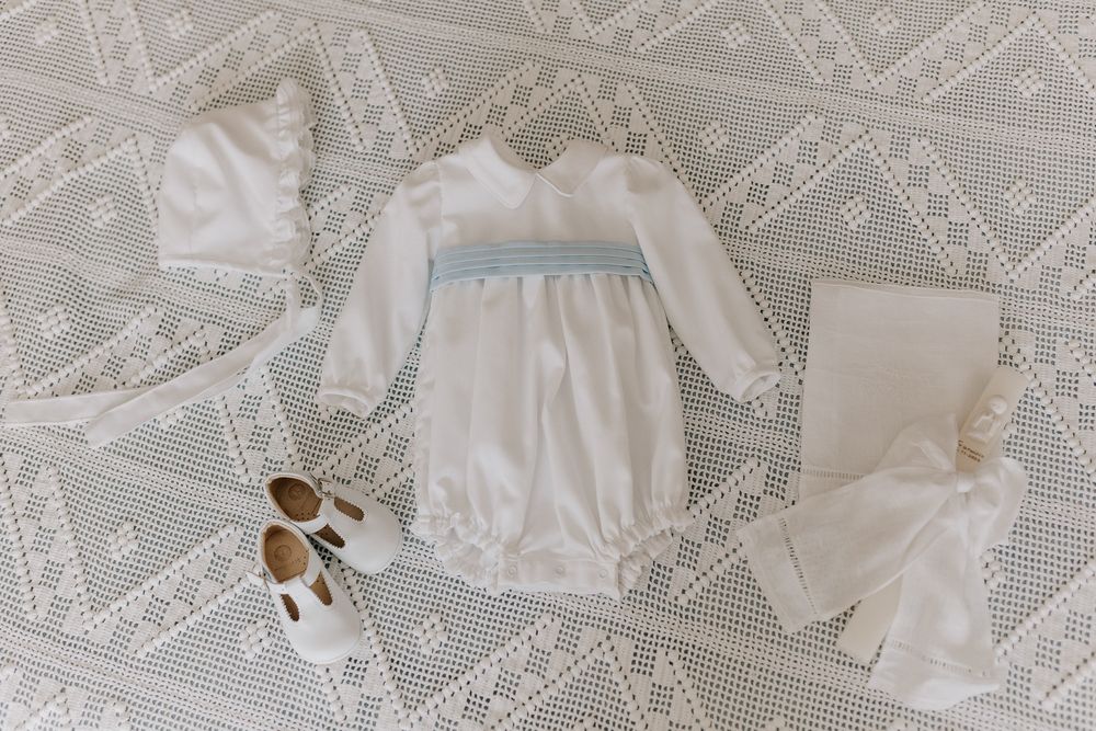 White baby christening outfit set with blue smocking, bonnet, booties, and pants on lace background.