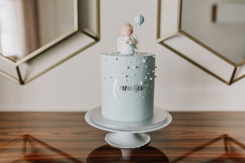 Light blue celebration cake with pearl details, baby figurine topper, and balloon on a white cake stand.