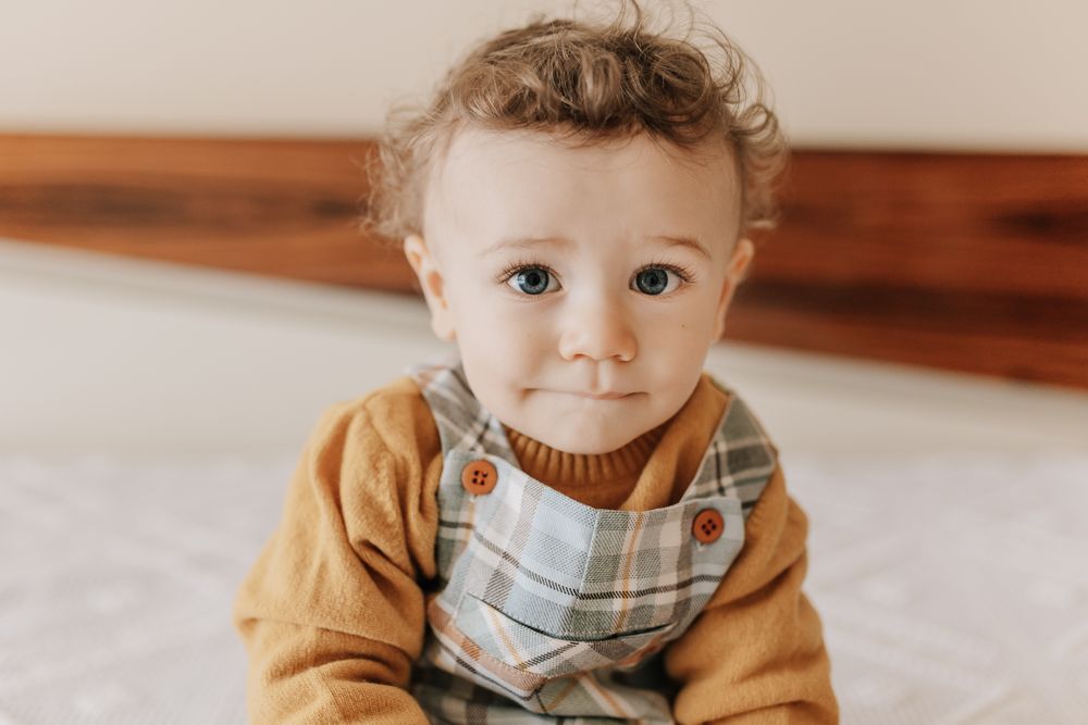 Adorable curly-haired baby in mustard sweater and plaid bib sits on white surface looking at camera.