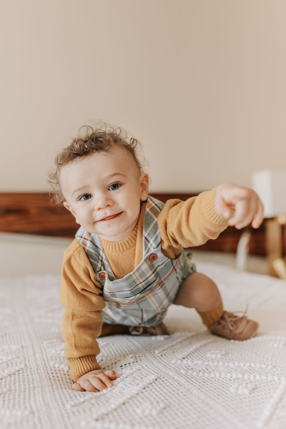 Adorable baby in mustard yellow sweater and plaid overalls crawling on white knit blanket, pointing forward.