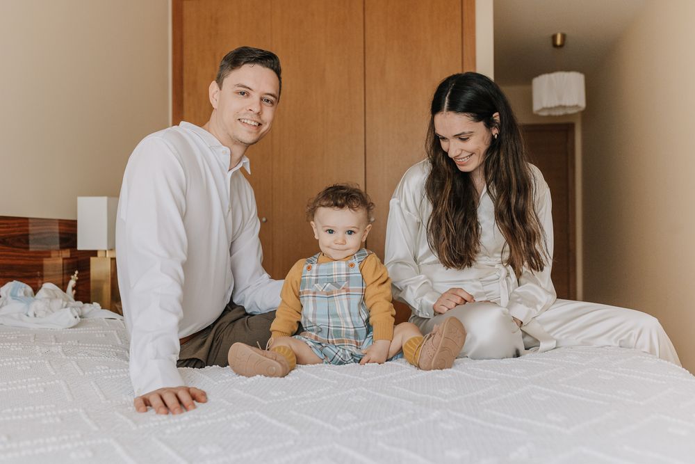 Happy family with parents and baby sitting together on a white bed smiling at camera.