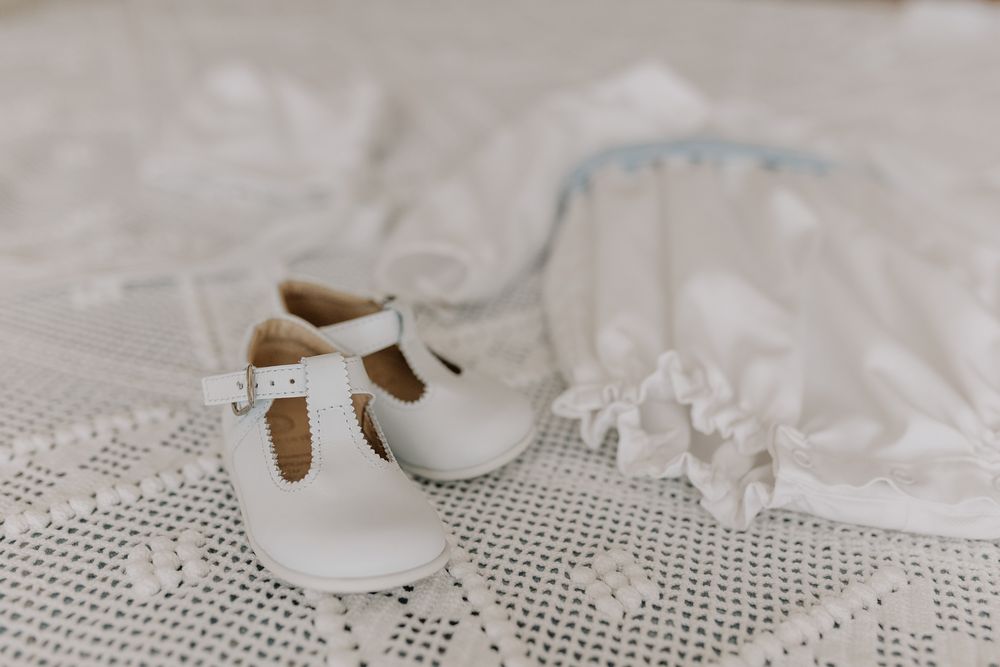 White leather baby T-bar shoes beside a delicate lace christening gown on a crochet blanket.