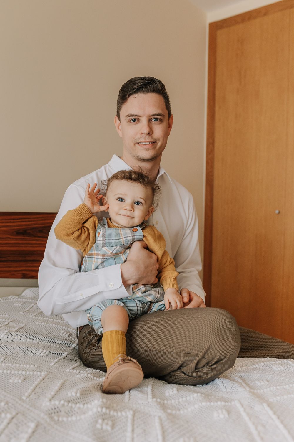 A smiling man in a white shirt sits cross-legged on a bed holding an adorable baby in plaid overalls and yellow socks.