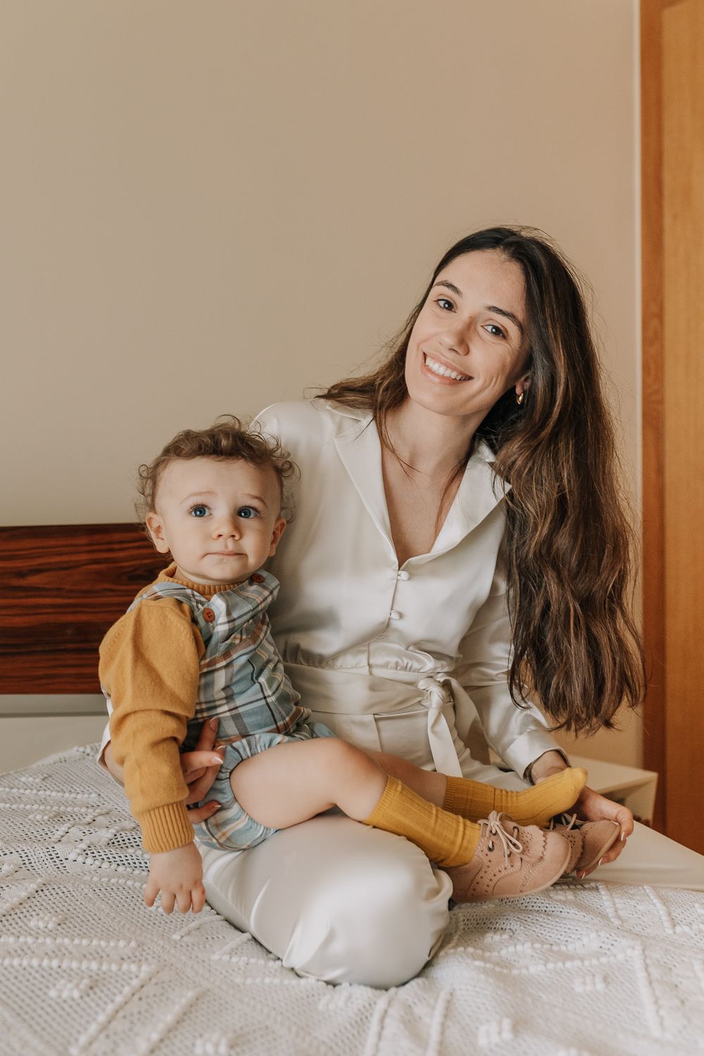 Smiling young mother sitting on bed with her baby toddler dressed in mustard yellow outfit in cozy home setting.