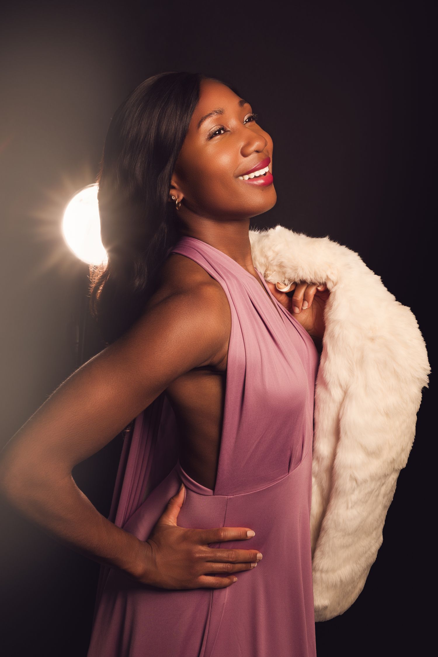 Elegant woman in red dress with pearl necklace and fur stole poses dramatically against dark background with bright light.