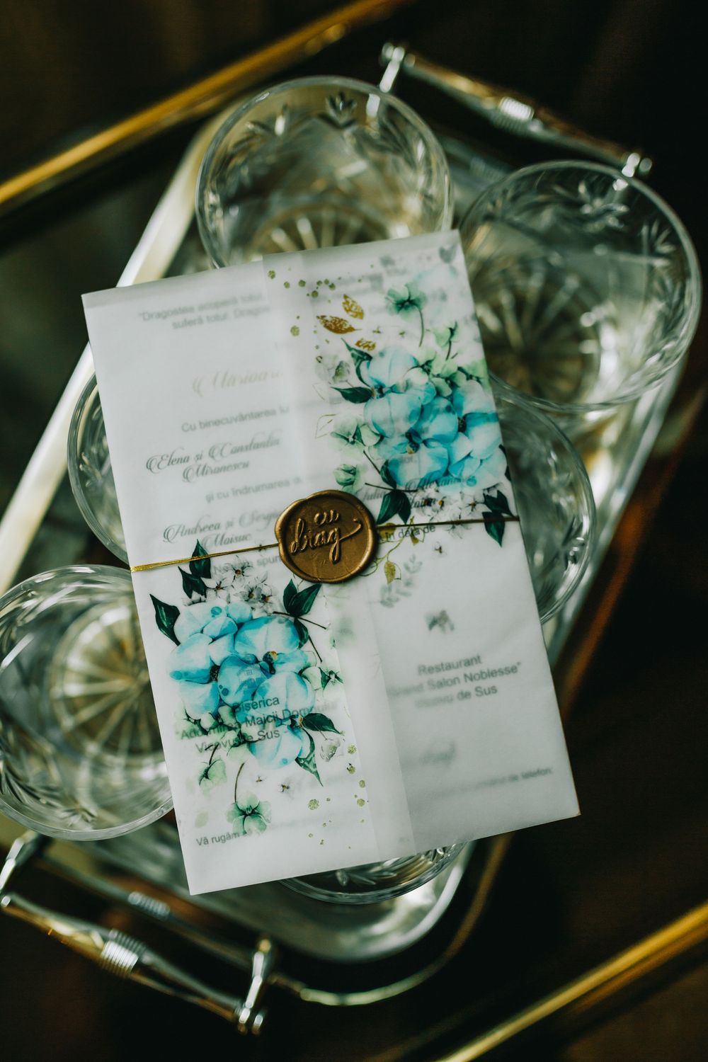 Wedding invitation with blue floral design and gold wax seal on crystal glasses tray.