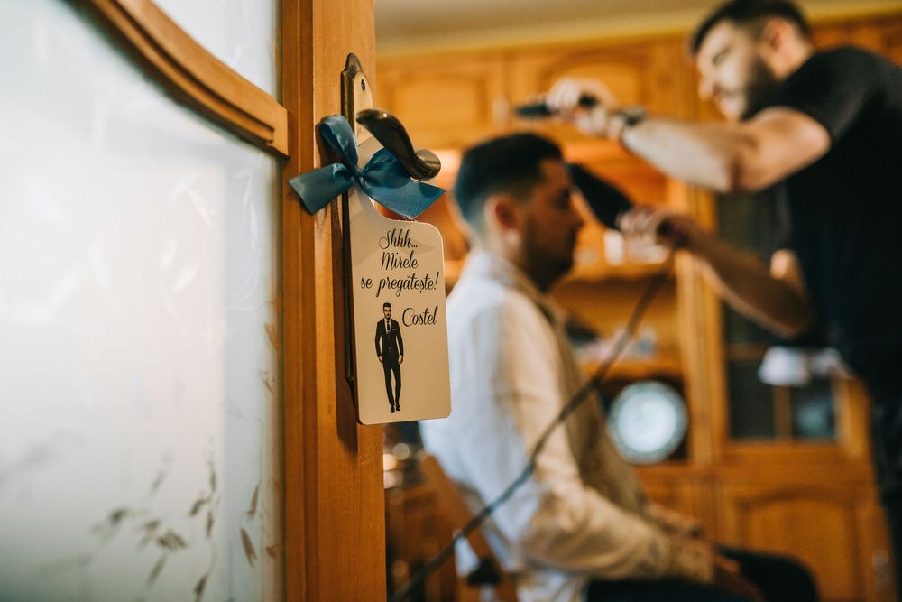 Groom getting hair styled before wedding with decorative door sign in foreground.
