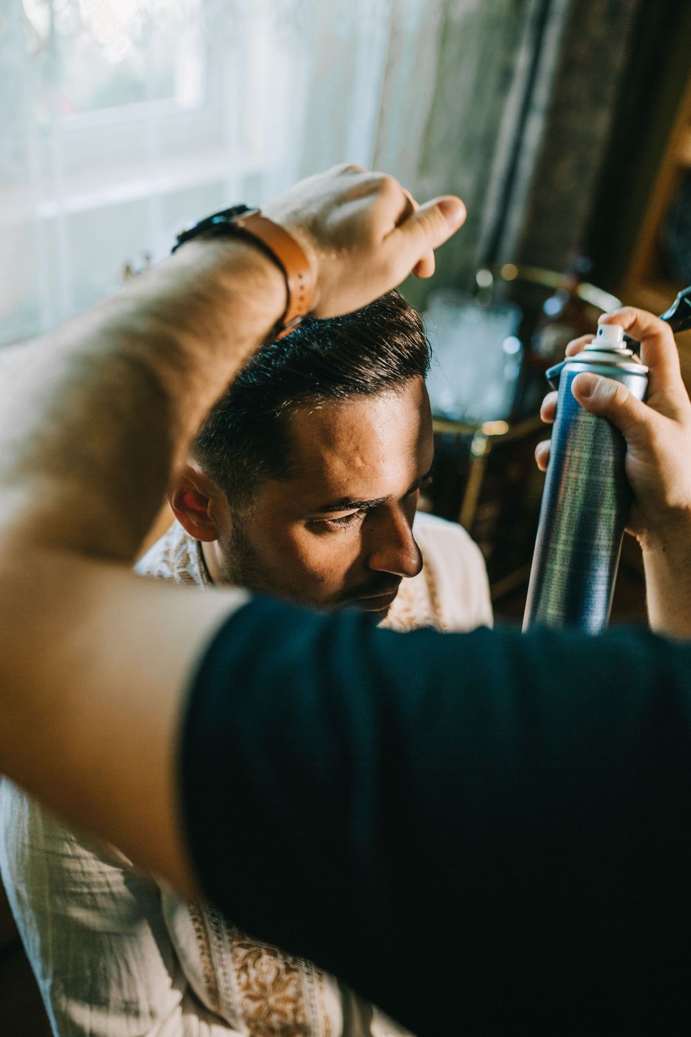 A hairstylist applying hairspray to a groom's hair during wedding preparations.