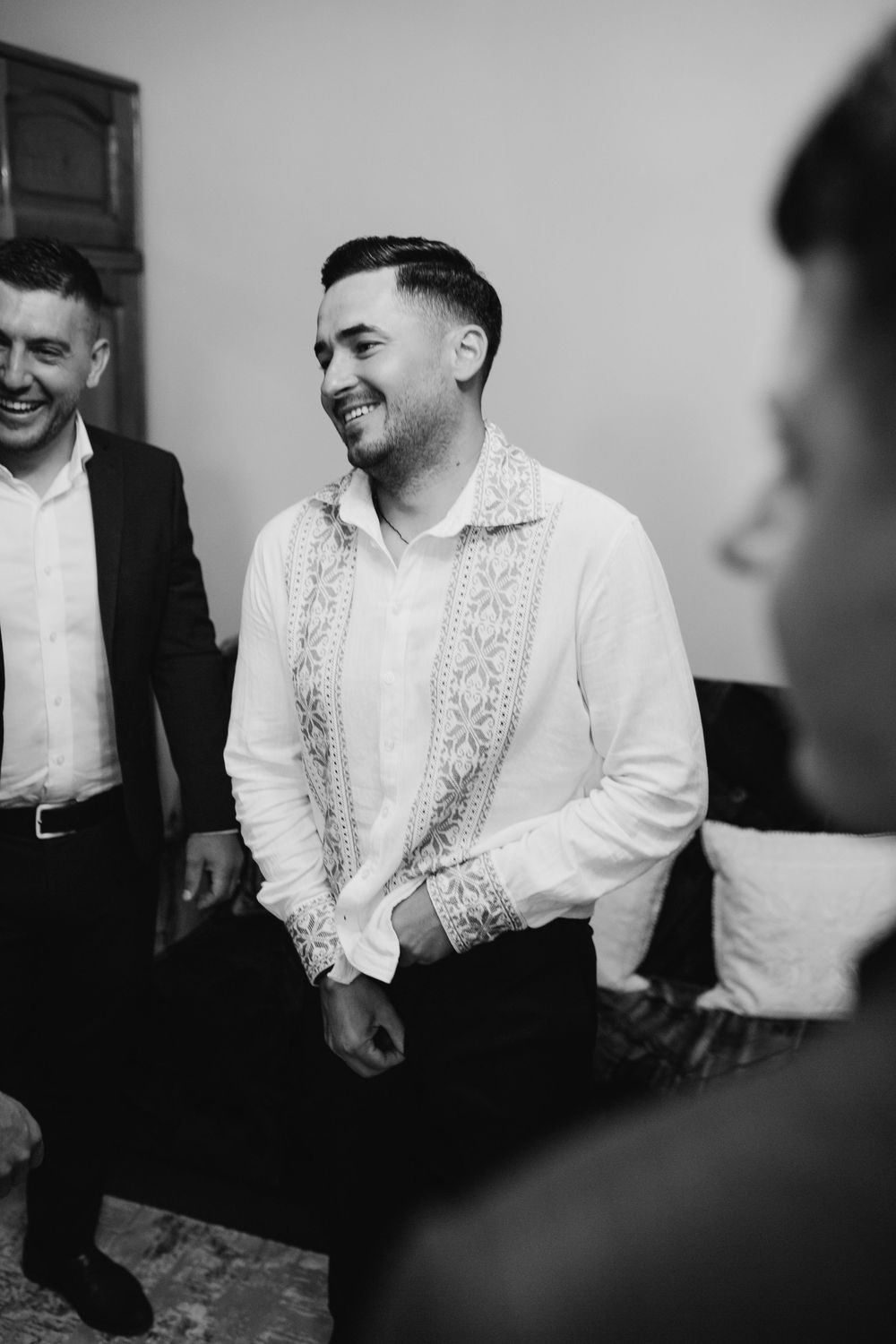 A smiling groom in an embroidered white shirt laughing with groomsmen in black and white photo.