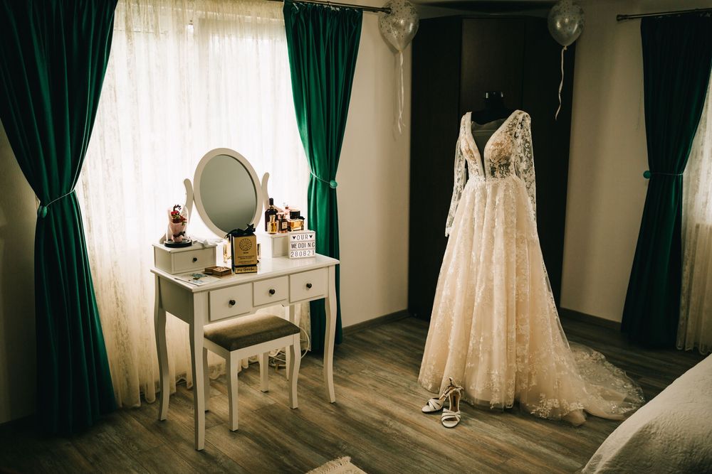 Elegant wedding dress on mannequin beside white vanity mirror with green curtains in bridal preparation room.