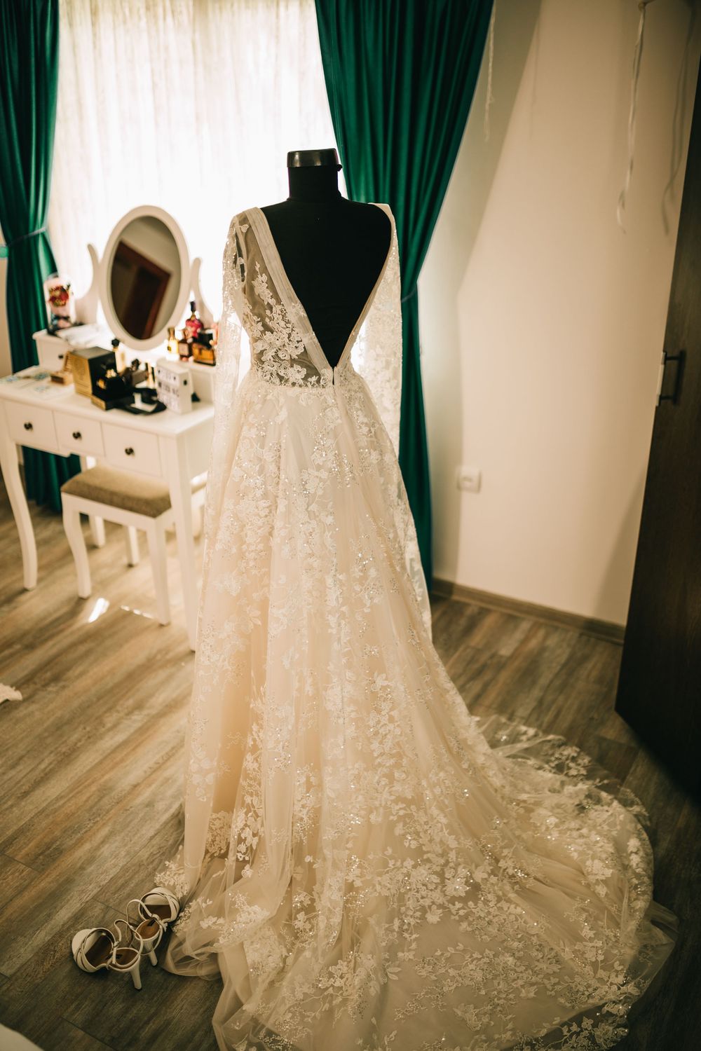 Stunning backless lace wedding gown with long train displayed on mannequin near window with teal curtains.