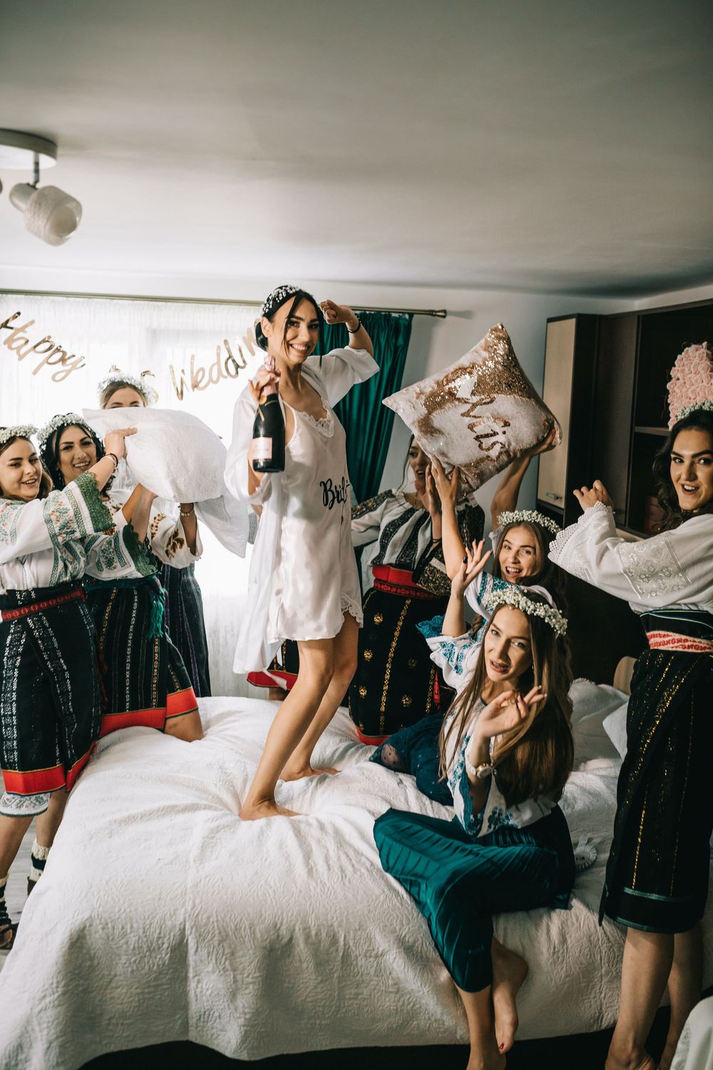 Excited bride in white robe celebrating with bridesmaids in traditional folk costumes on bed with champagne.