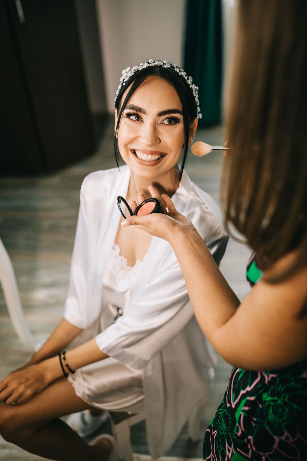 Smiling bride in white robe getting makeup applied by artist holding brushes and powder palette.