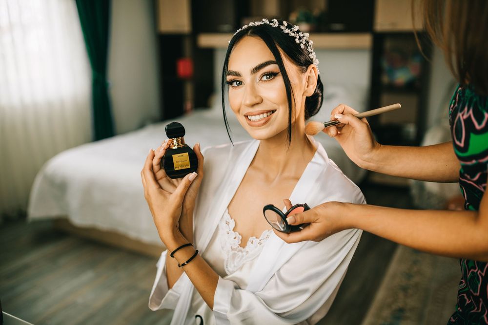 Beautiful bride in white lace robe holding perfume bottle while makeup artist applies blush.