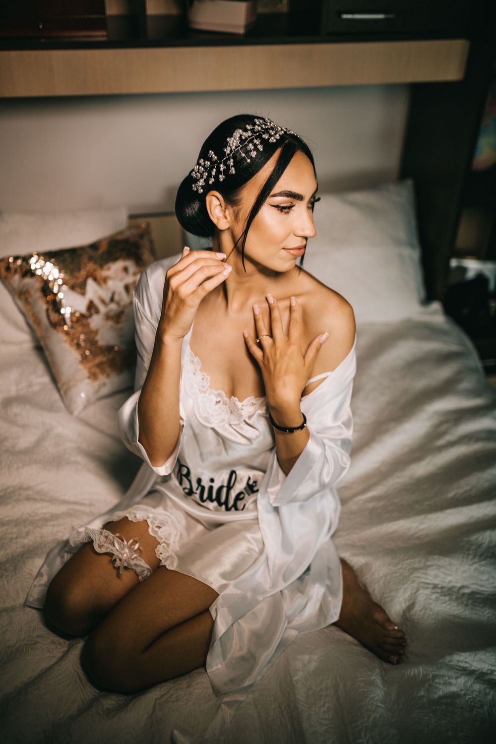 Elegant bride in white lace lingerie and robe sitting on bed adjusting earring with headpiece.