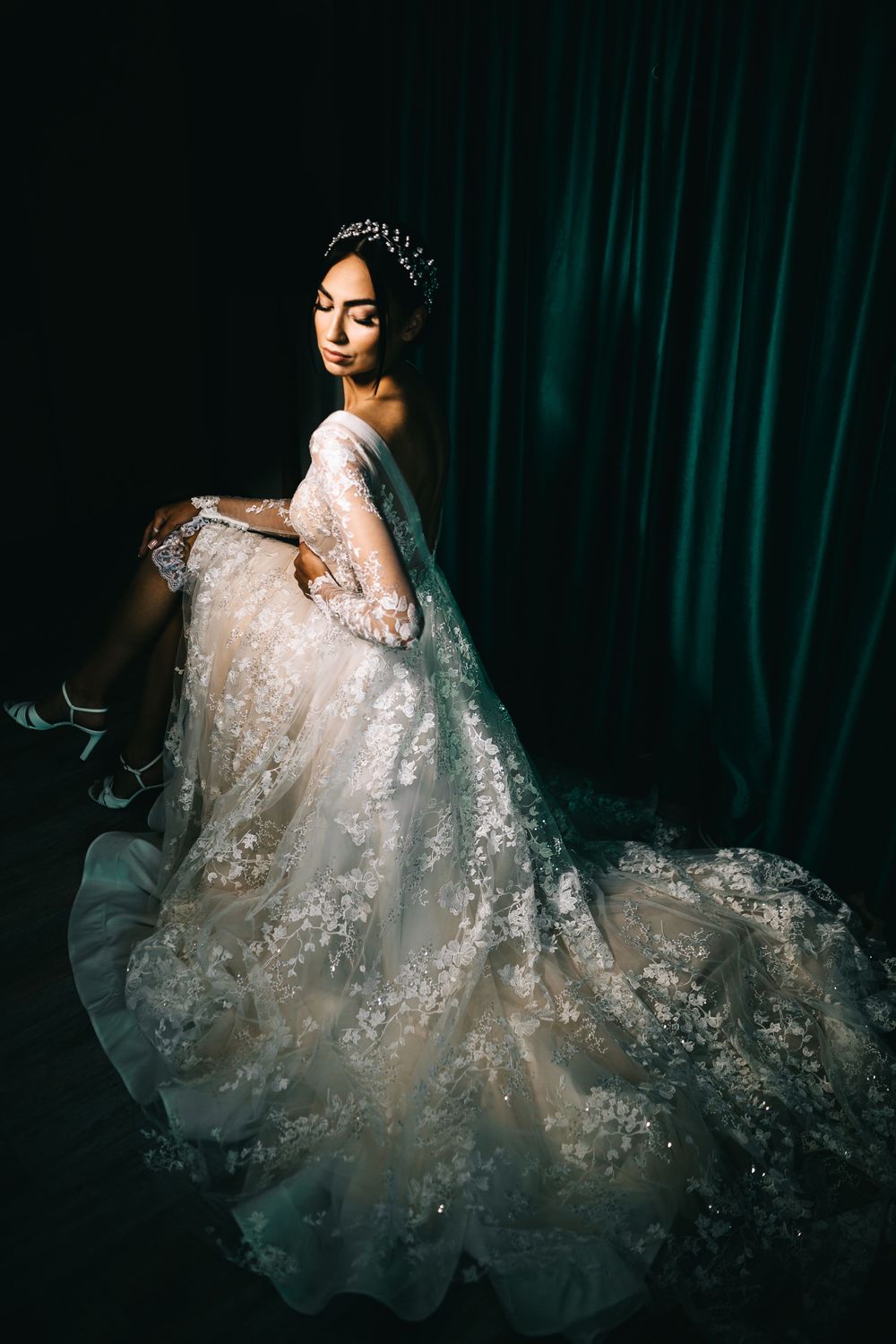 Dramatic portrait of bride in ornate lace wedding dress seated against dark green curtain backdrop.