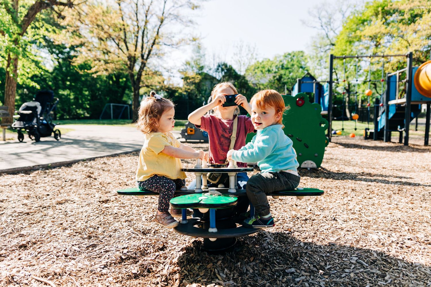 Three toddlers playing on a spinning merry-go-round at a sunny playground surrounded by trees.
