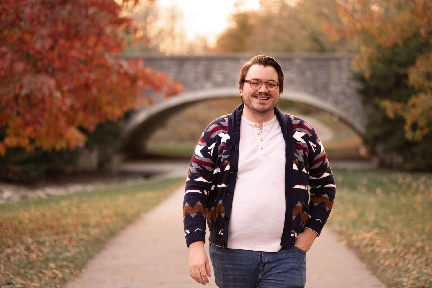 Man smiling in patterned jacket on autumn park path with stone bridge and colorful foliage in background.