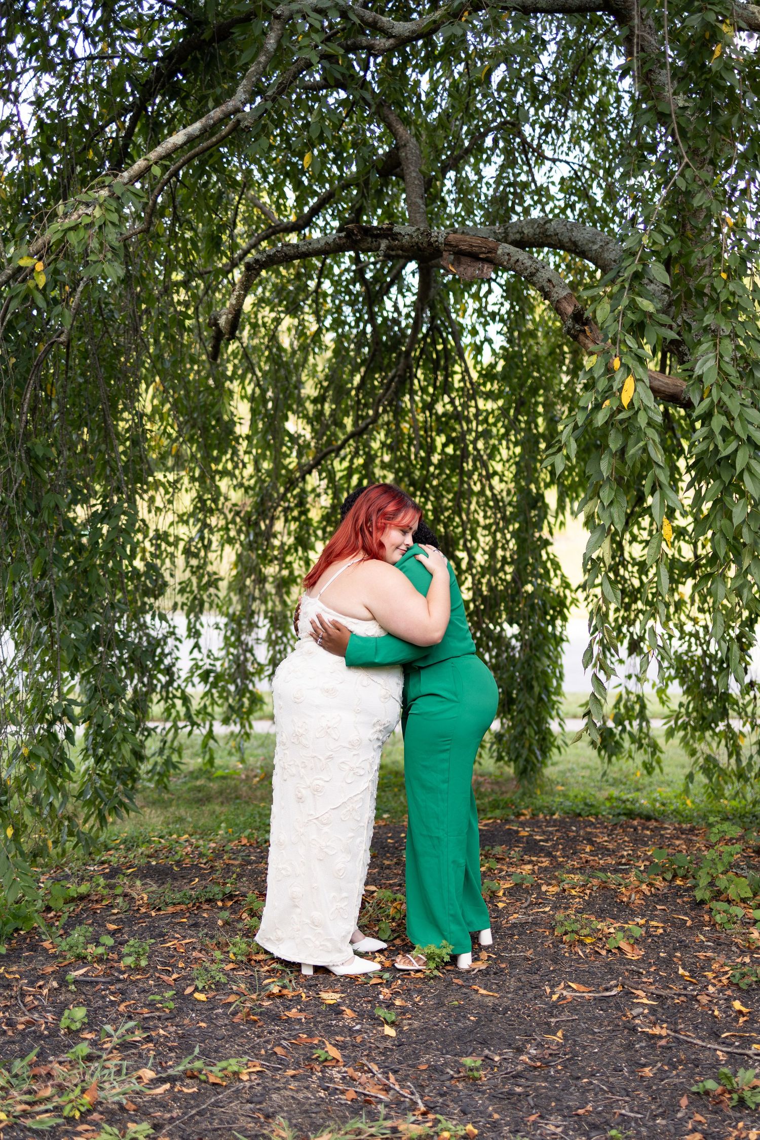 Two women in formal gowns embrace under a weeping cherry tree in Ault Park.