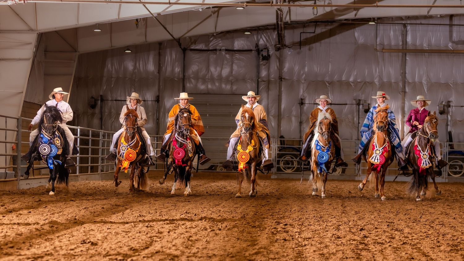 Seven riders on decorated horses perform in an indoor rodeo arena, wearing colorful western attire.