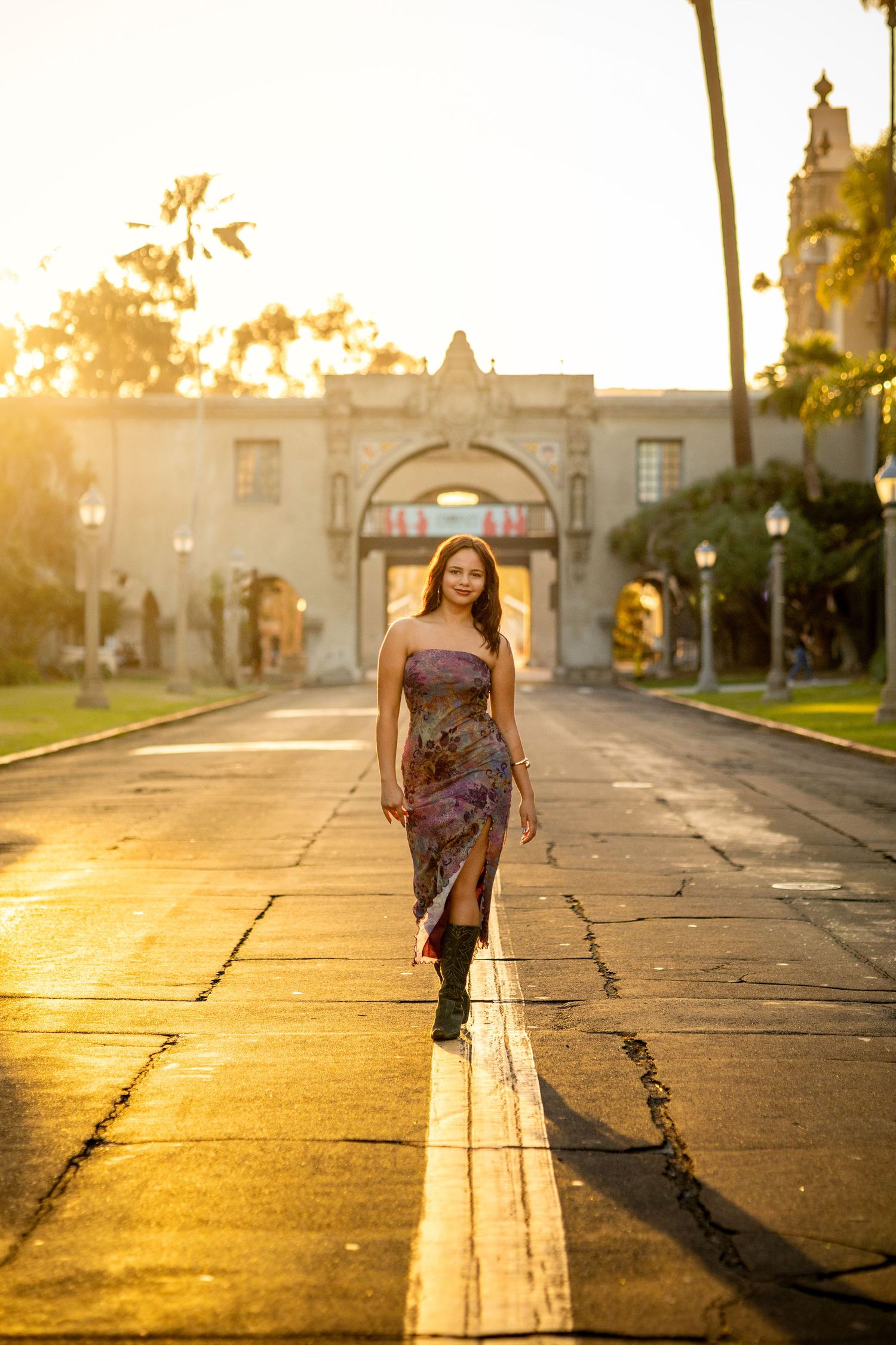 A high school senior in black shirt and jeans sits outdoors at sunset with golden bokeh lighting in the background.