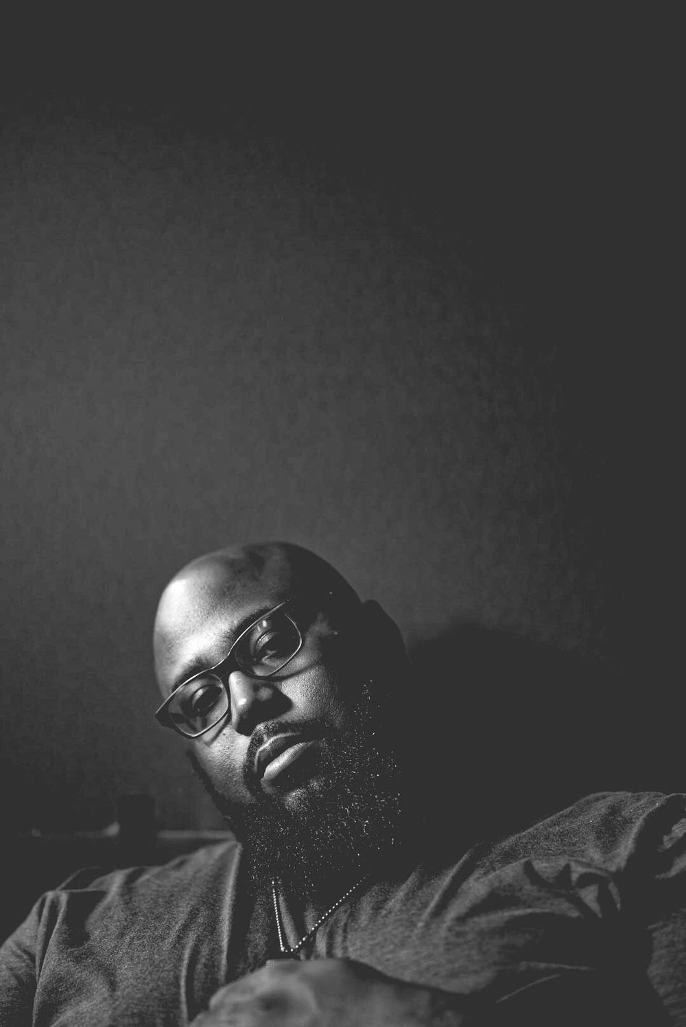 Bald Black man with glasses and beard in low-key black and white portrait, looking down.