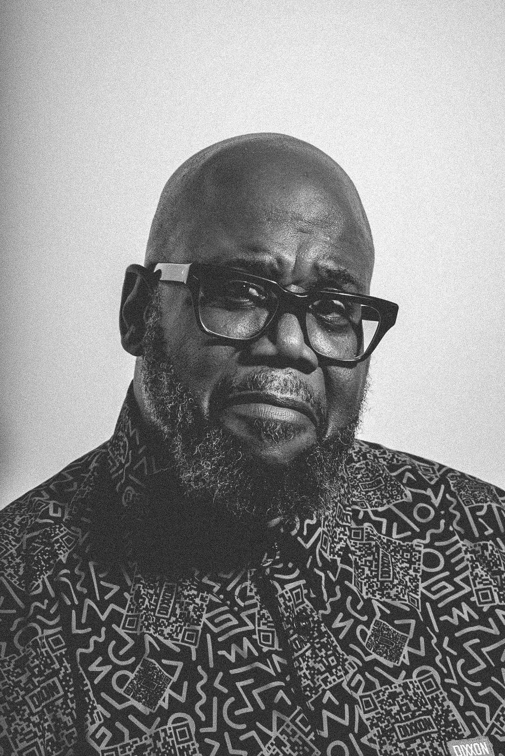Bald Black man with beard and glasses wearing patterned shirt in black and white portrait.