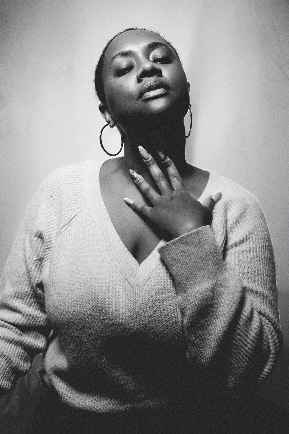 Black and white portrait of a woman in a knit sweater with hoop earrings, eyes closed, hand on chest.