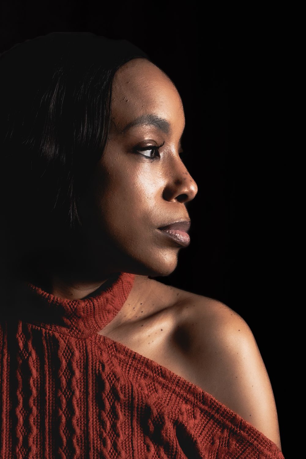 A Black woman in a rust-colored knit top poses in dramatic side profile lighting against a dark background.