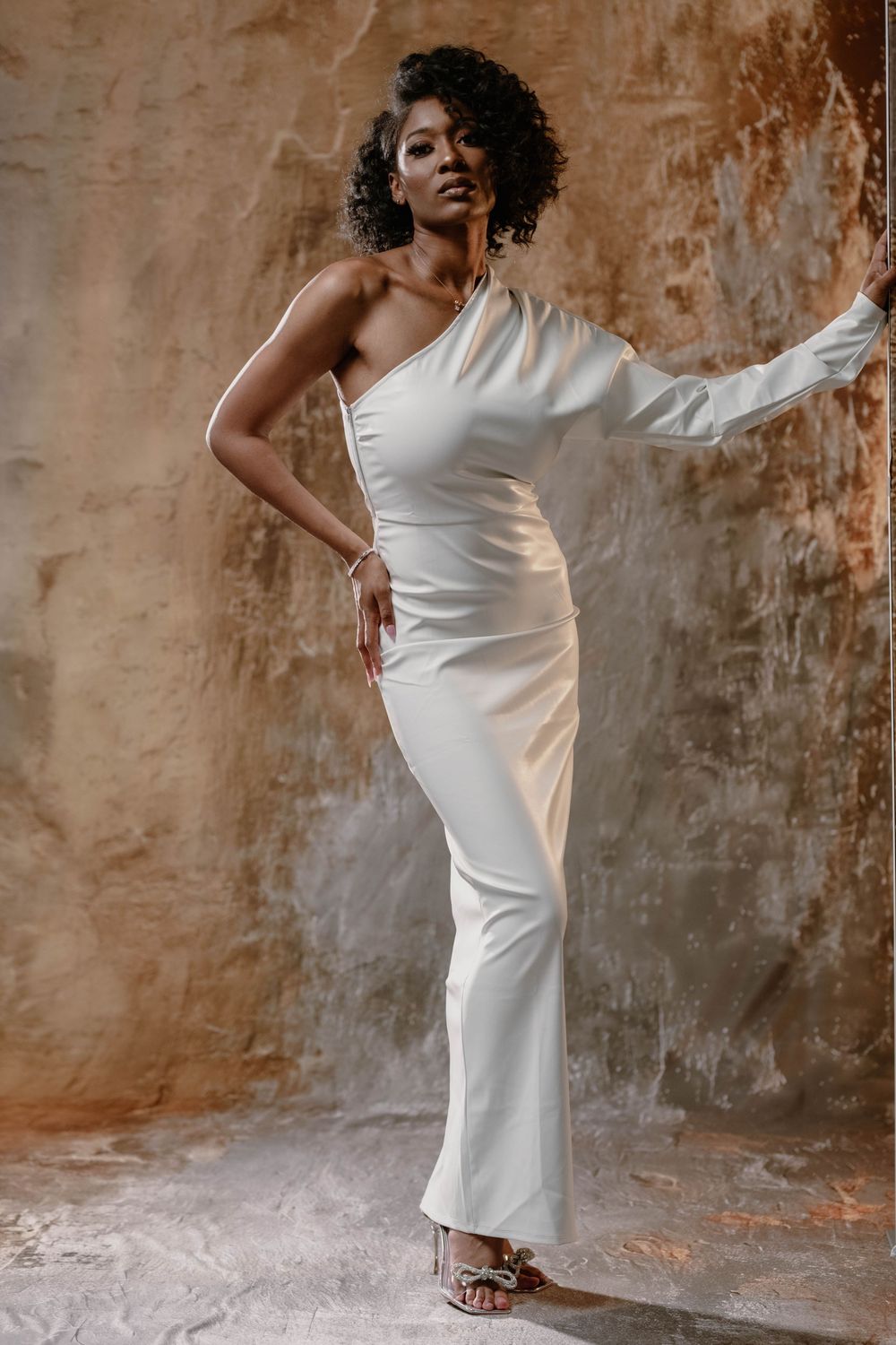 Elegant Black woman in white one-shoulder gown posing against textured wall in studio.