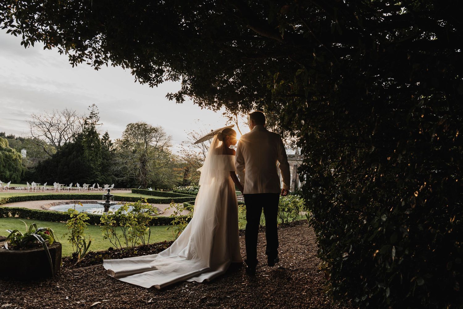 Bride and groom stand hand in hand in a sunlit garden, framed by lush greenery, with a fountain in the background.