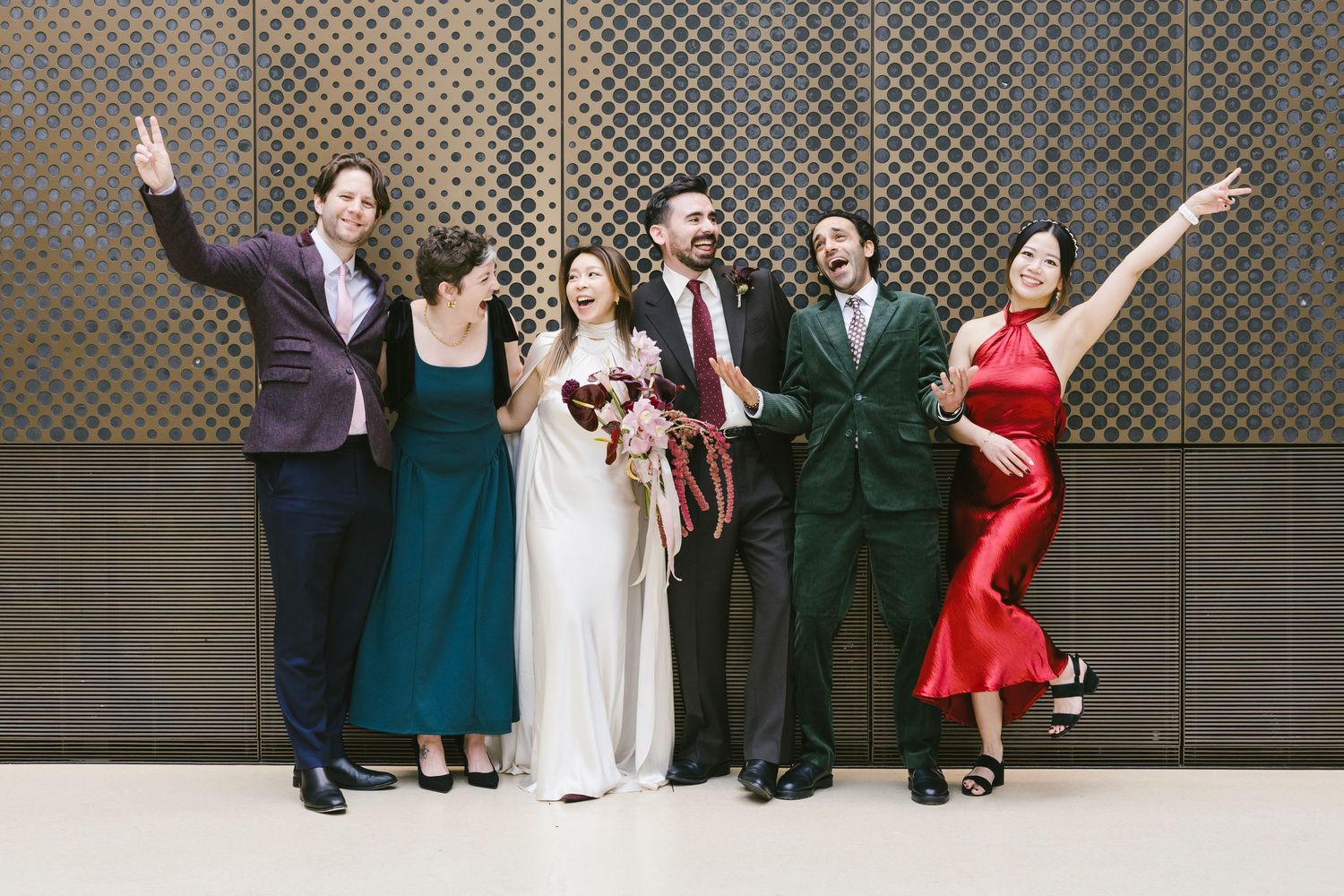 Elegant couple walking through a grand hall, groom in dark suit, bride in white pantsuit holding a bouquet.