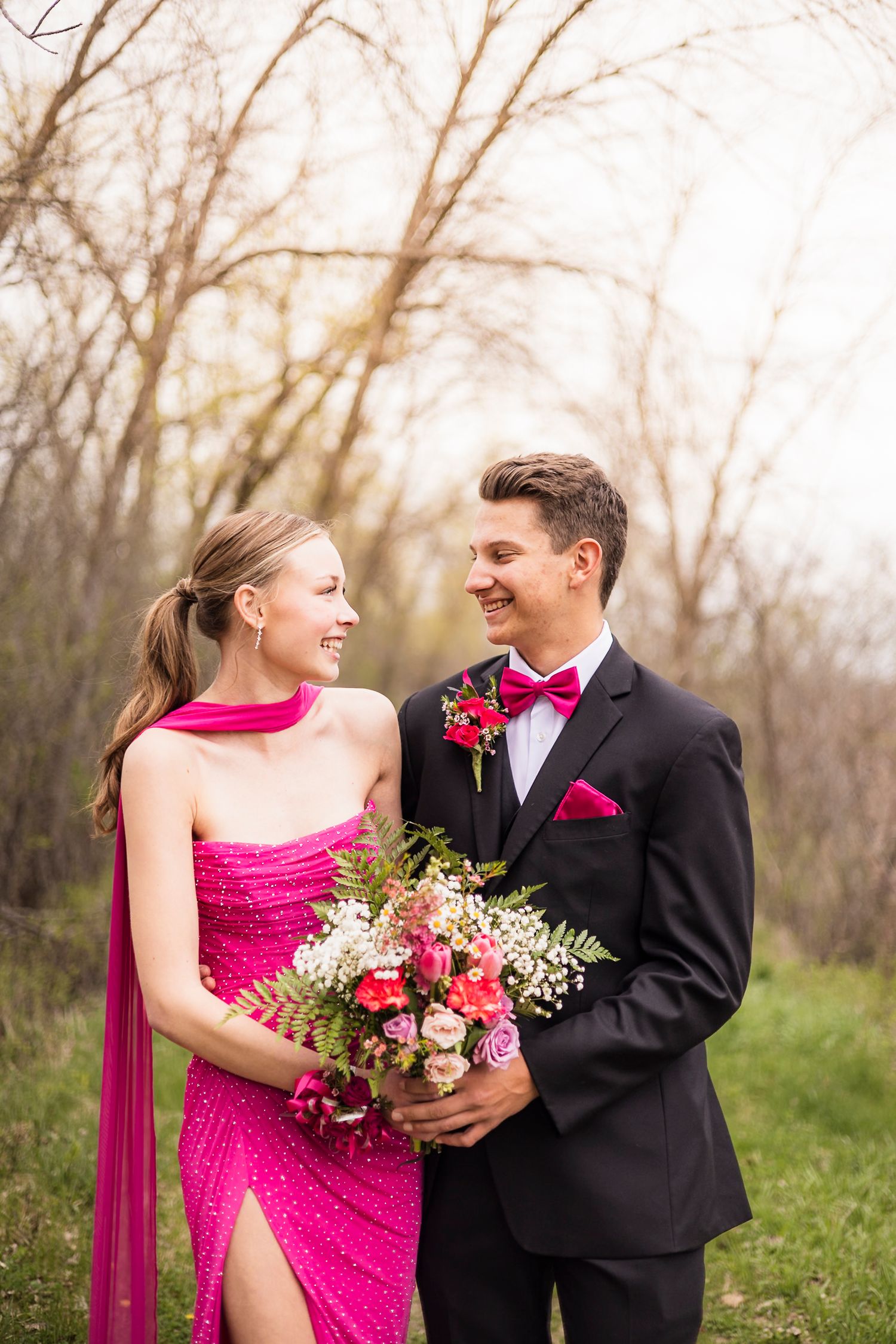 Couple in matching pink and black prom attire smiling at each other outdoors, holding a bouquet of pink and white flowers.