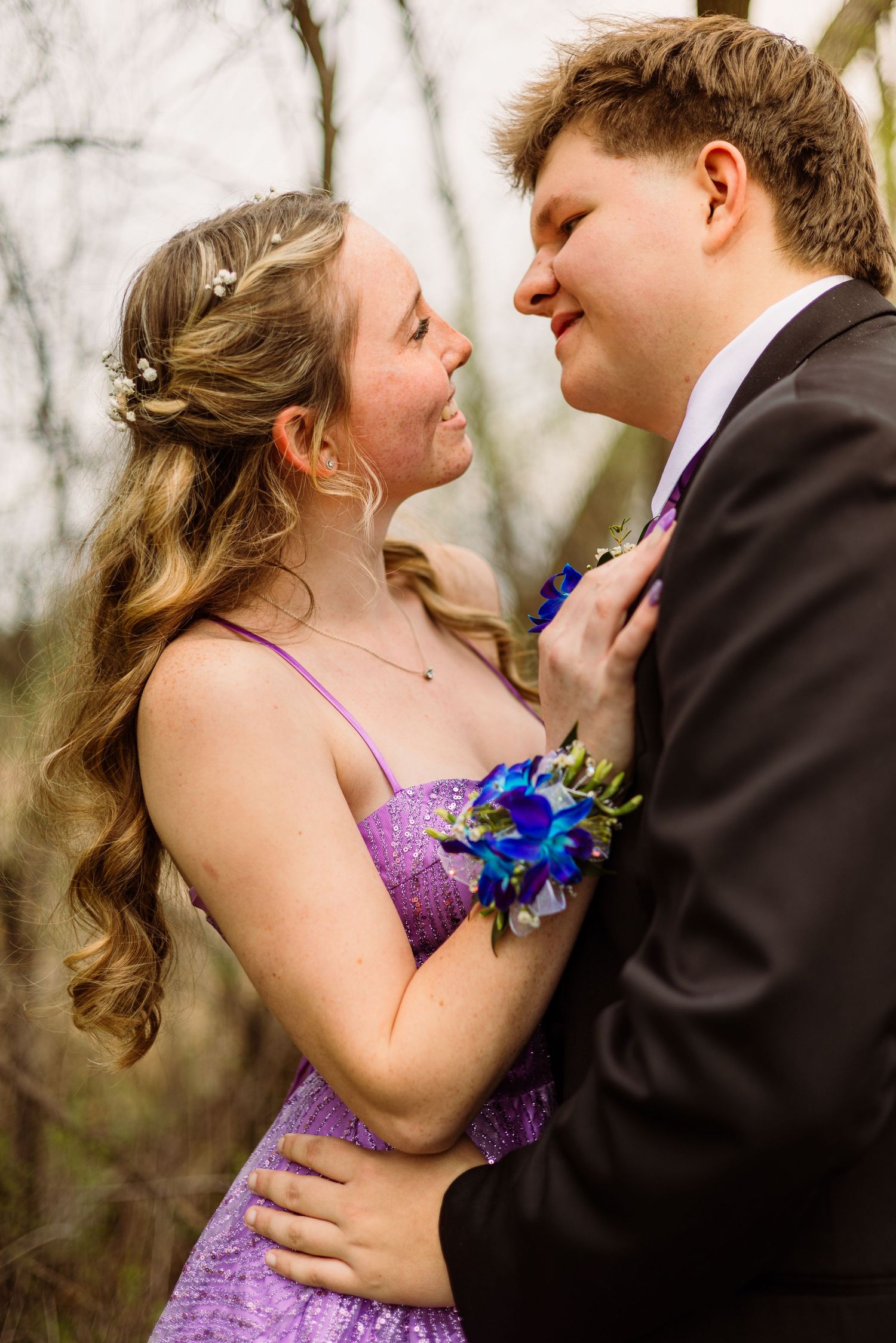 Young couple in prom attire, girl in purple sequin dress with blue corsage, embracing and smiling at each other outdoors.