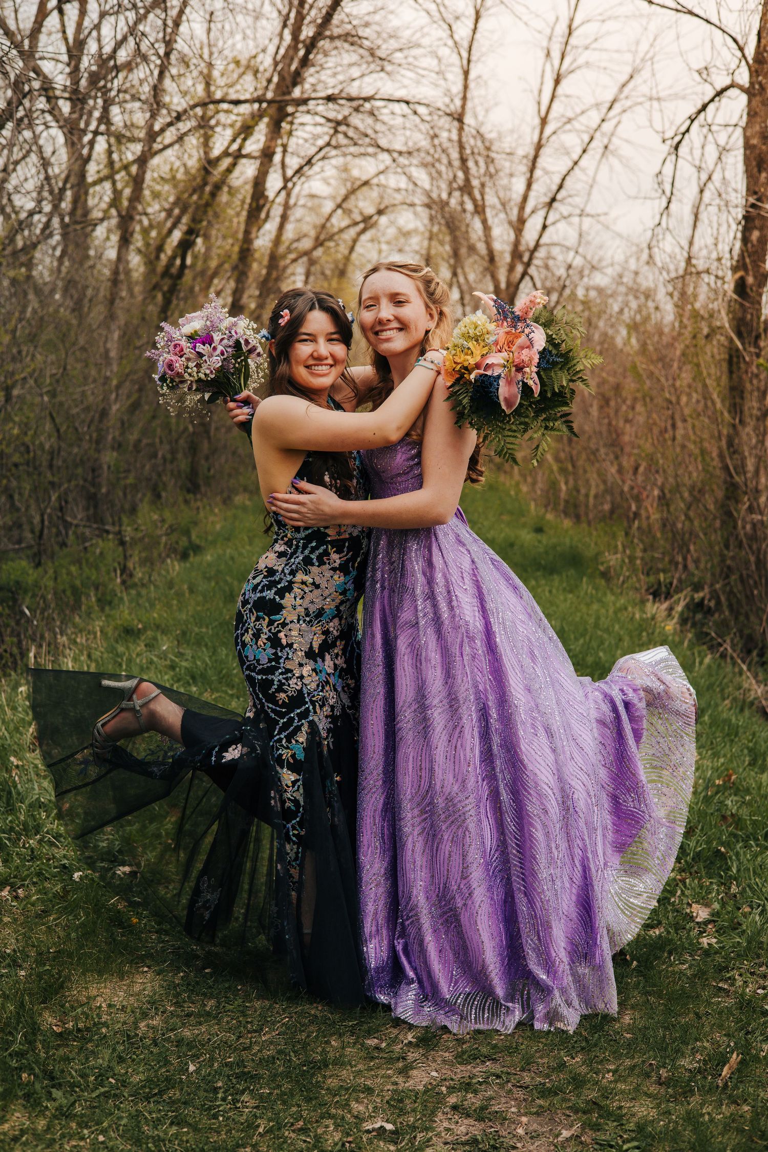 Two young women in formal gowns holding bouquets embrace outdoors among bare trees in spring.
