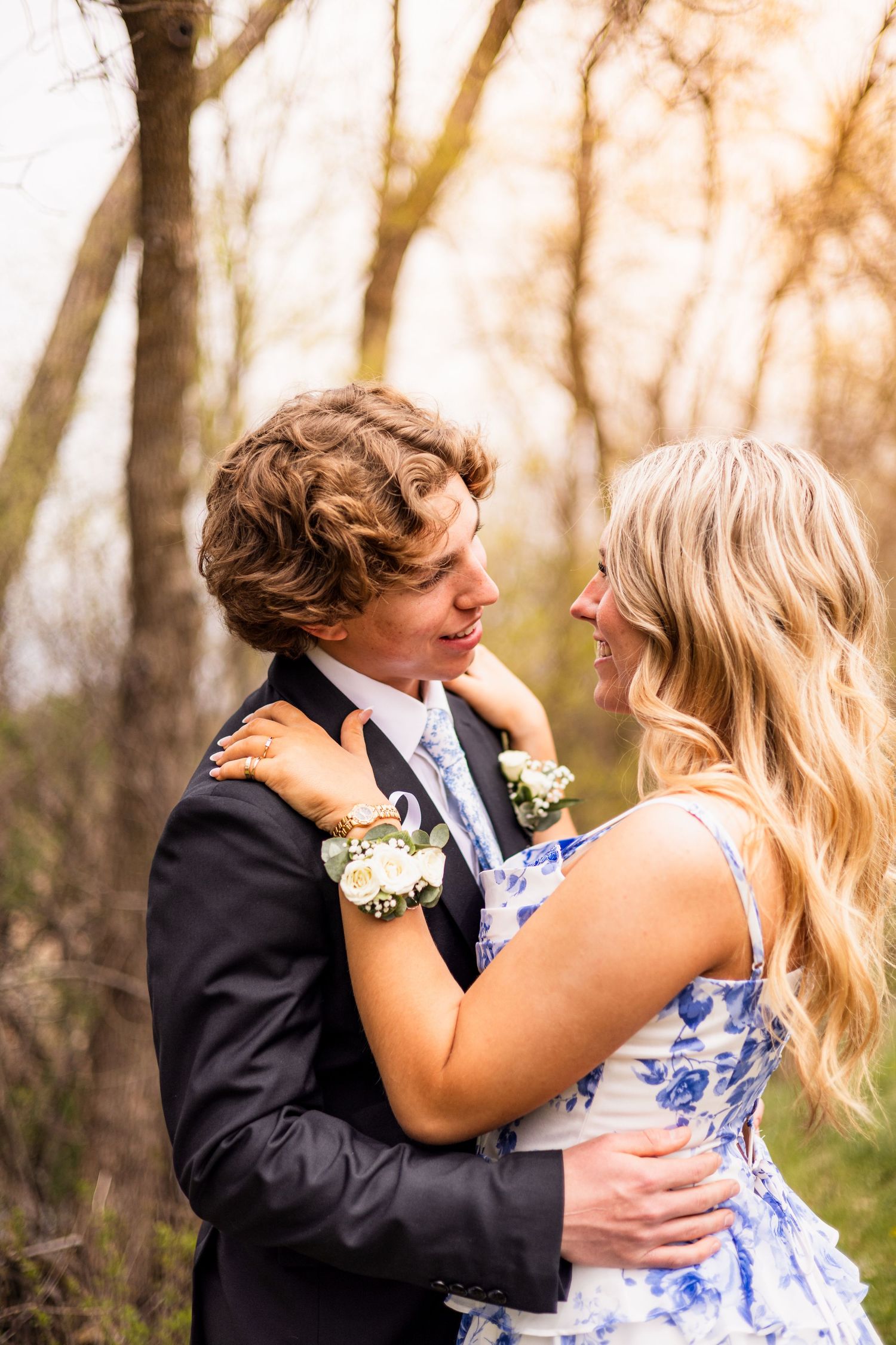 A young couple in prom attire embrace outdoors, the girl wearing a floral dress and corsage, smiling at her date in a suit.