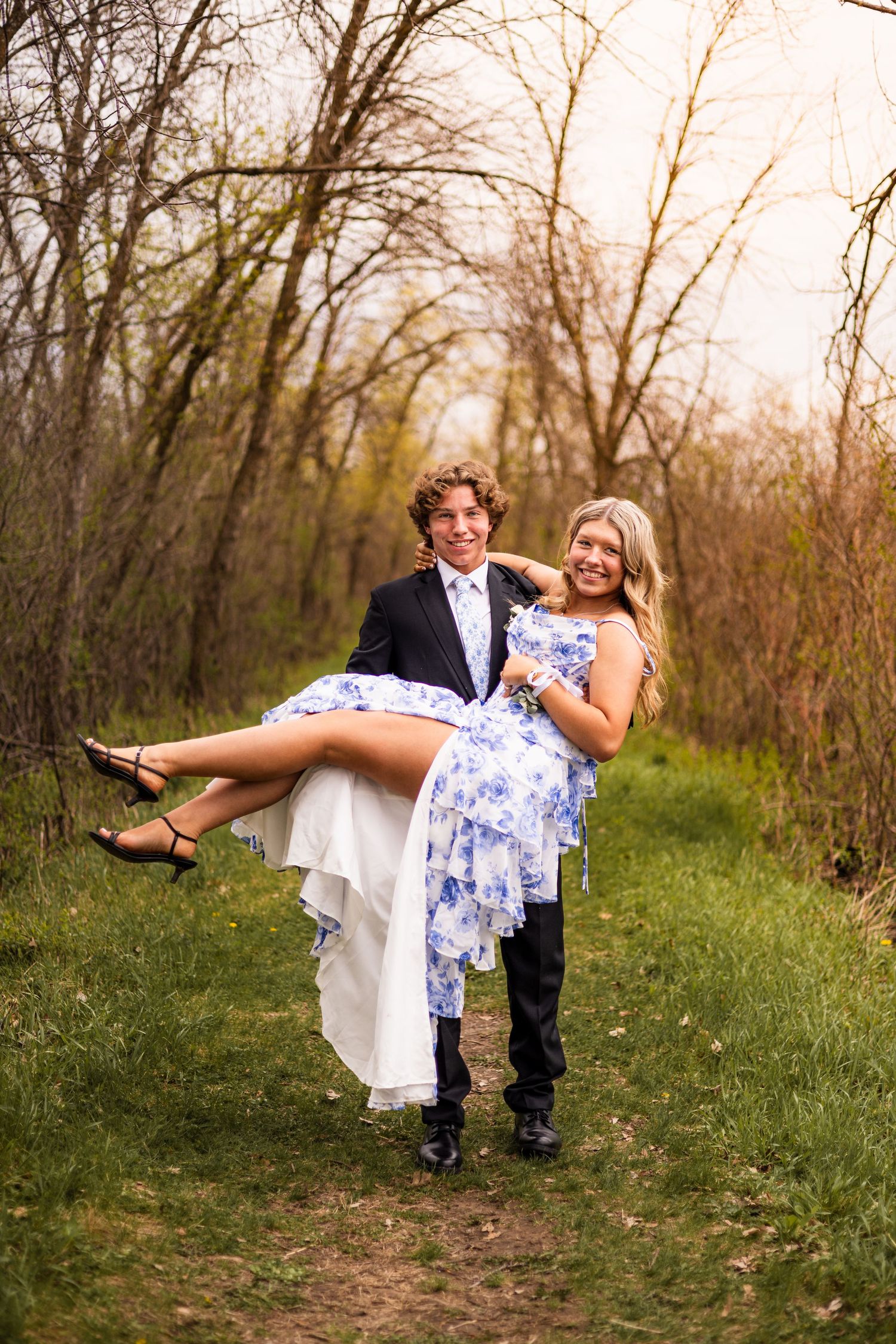 Young man in suit carrying smiling blonde woman in blue floral dress on a wooded path during prom photos.