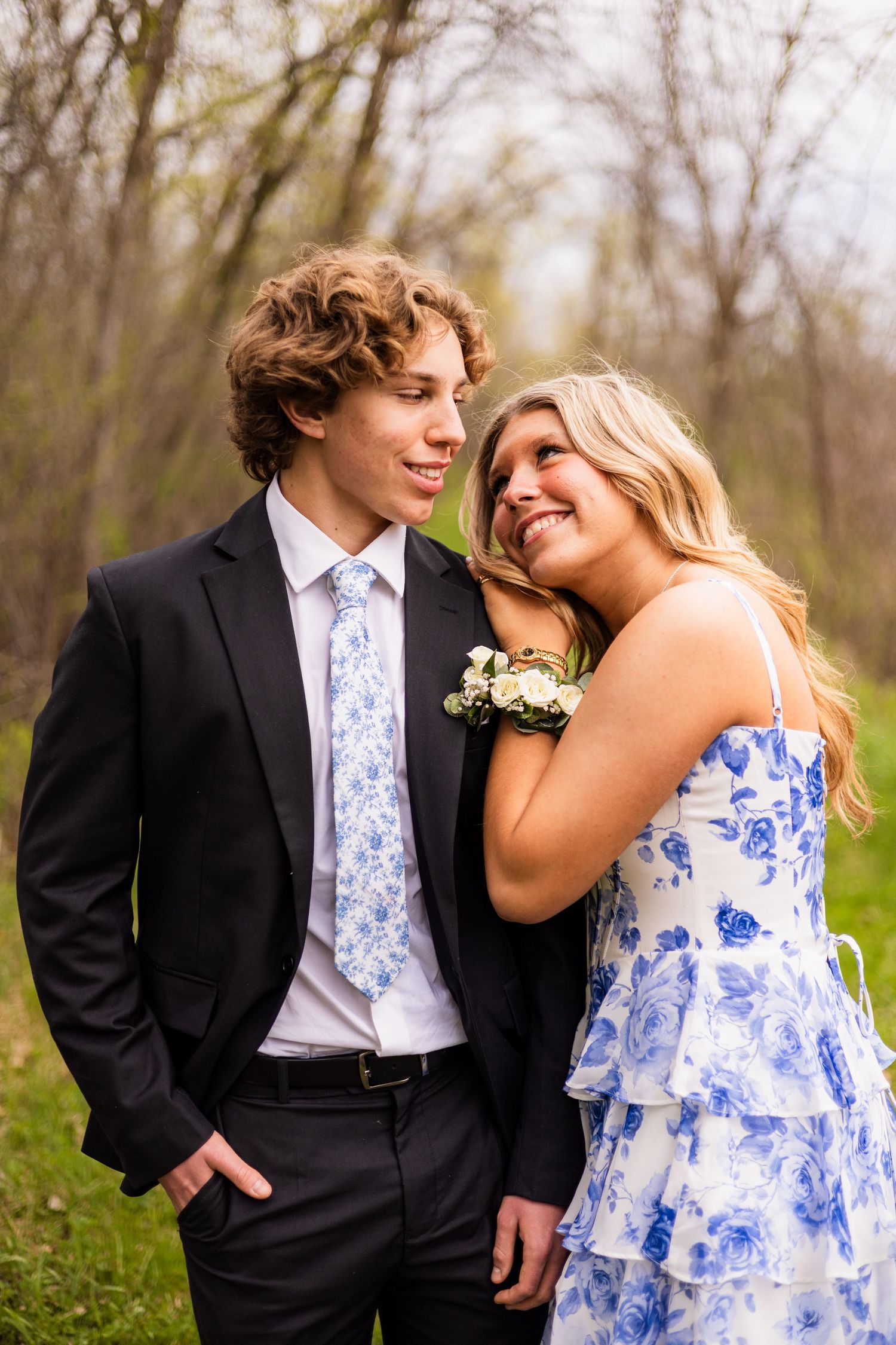 Young couple in matching blue floral outfits posing outdoors for prom photos, smiling together among spring trees.