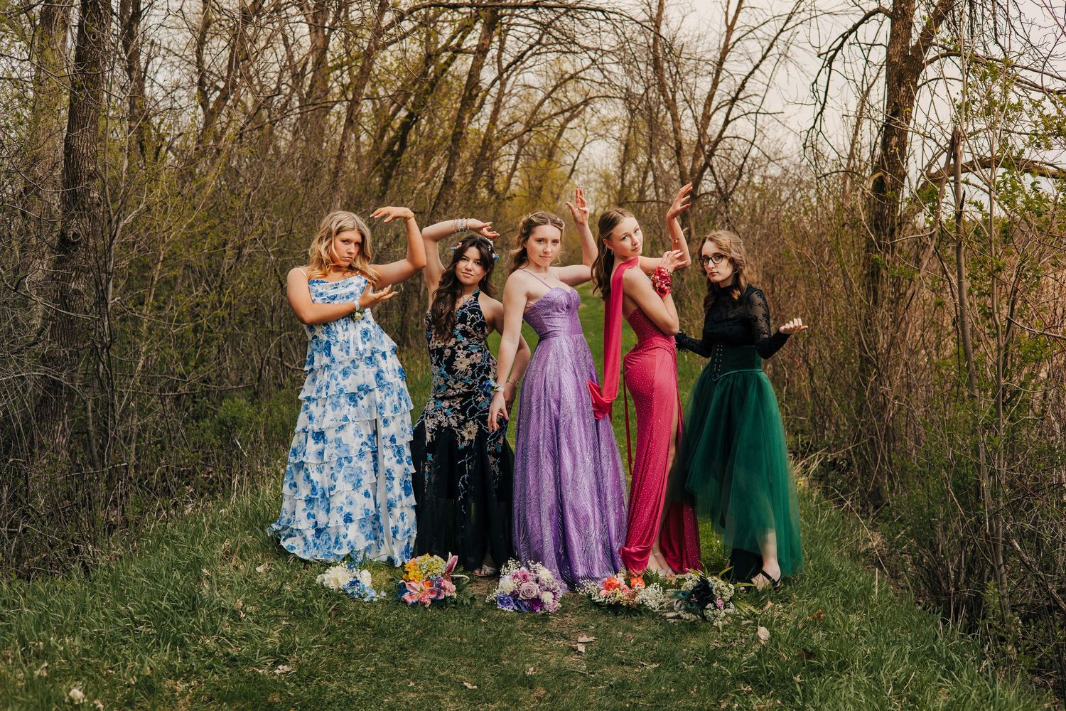 Five young women in colorful formal gowns posing outdoors among trees, dressed for prom or a special occasion.