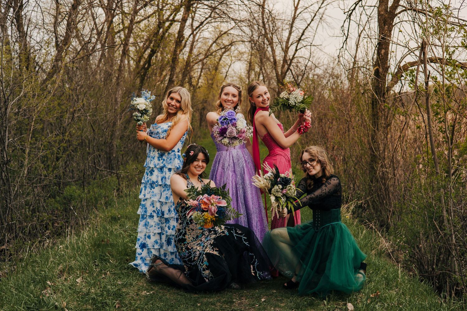 Five young women in colorful formal gowns pose outdoors holding flower bouquets among trees for a prom or special occasion.