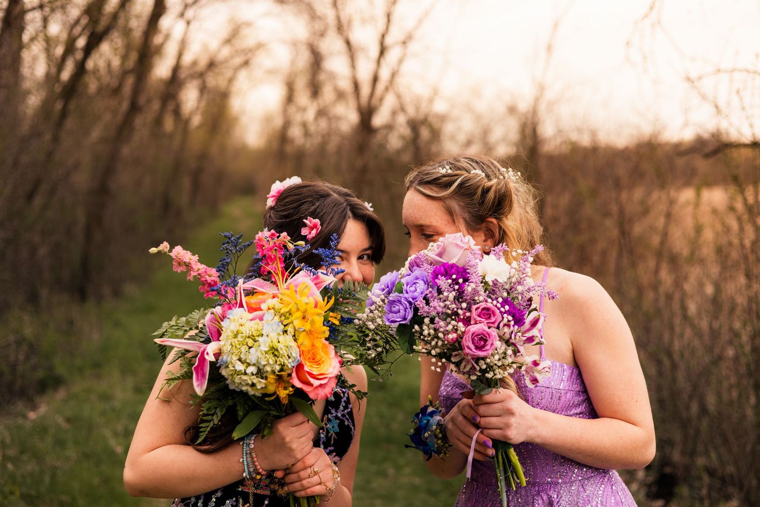 Two women smelling colorful wildflower bouquets outdoors in a wooded setting during golden hour.