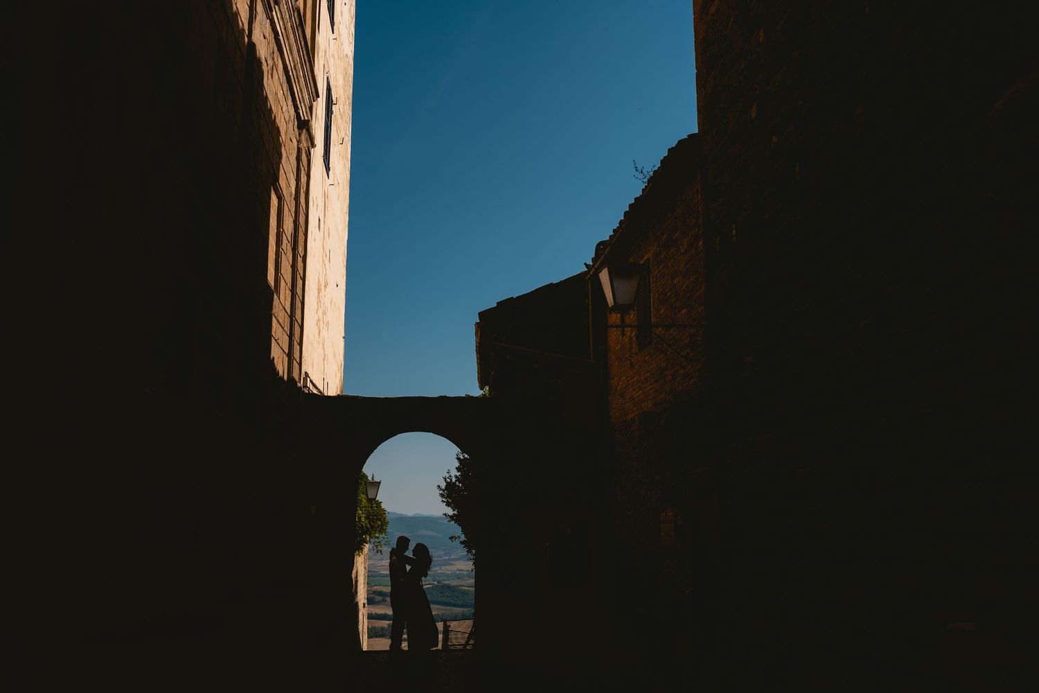 Silhouette of a couple embracing under a stone arch in ancient ruins, framed by blue sky.