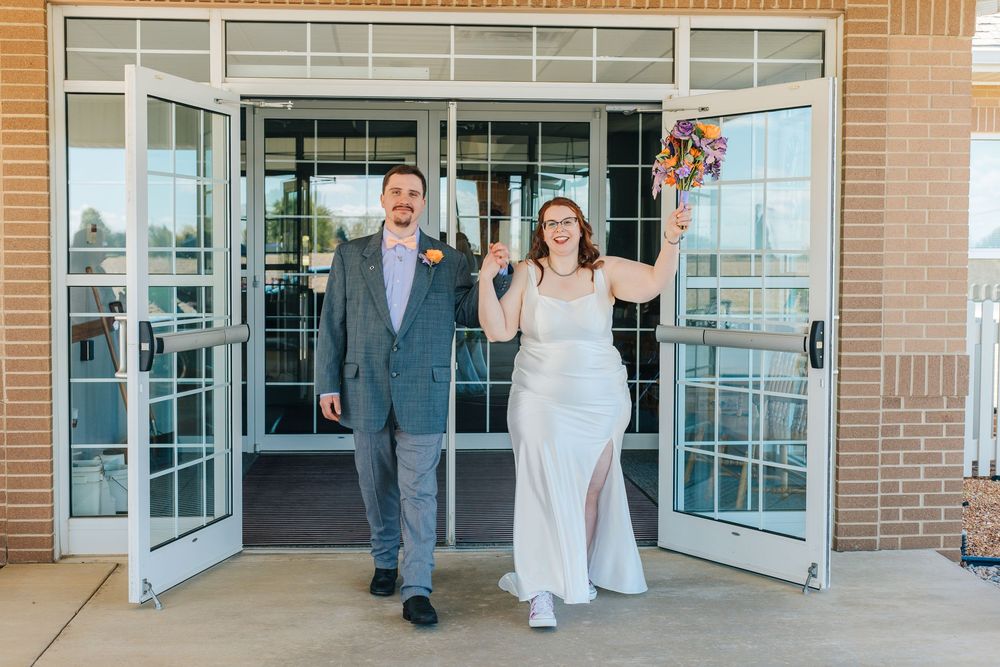 Joyful newlyweds exit wedding venue, bride in white gown holding colorful bouquet, groom in gray suit, brick building backdrop.