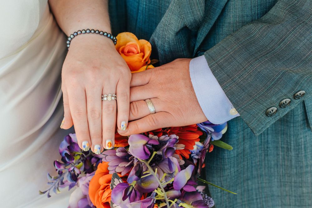 Close-up of newlyweds hands showing wedding rings resting on a colorful bridal bouquet of orange and purple flowers.